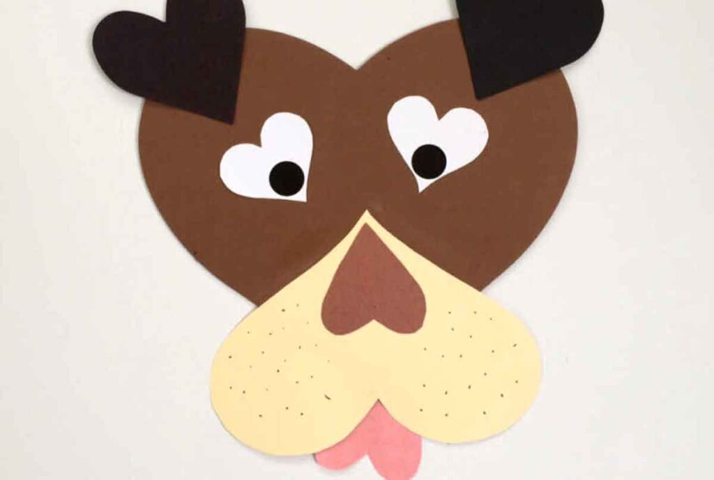 An image of a dog's face crafted from paper cutouts, perfect for Valentine paper crafts. The heart-shaped visage features brown and beige tones, black circle eyes, a heart-shaped nose, and a pink tongue, with two black heart shapes for ears.