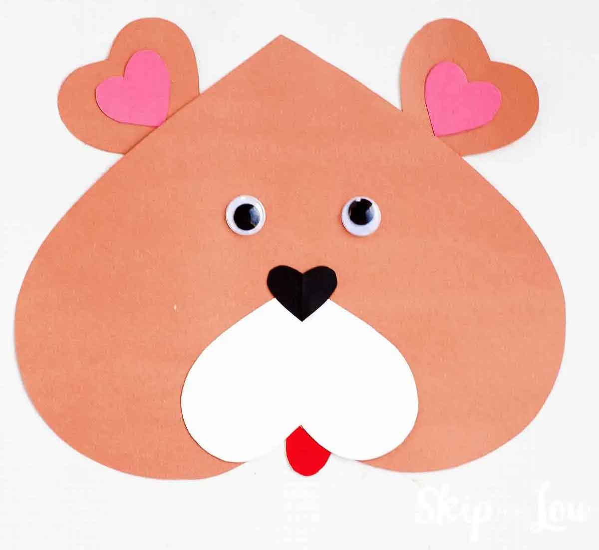 This charming Valentine paper craft features a heart-shaped brown bear's face, complete with googly eyes, a black nose, white cheeks, and a small red tongue. Pink heart shapes add a cute touch as inner ears.