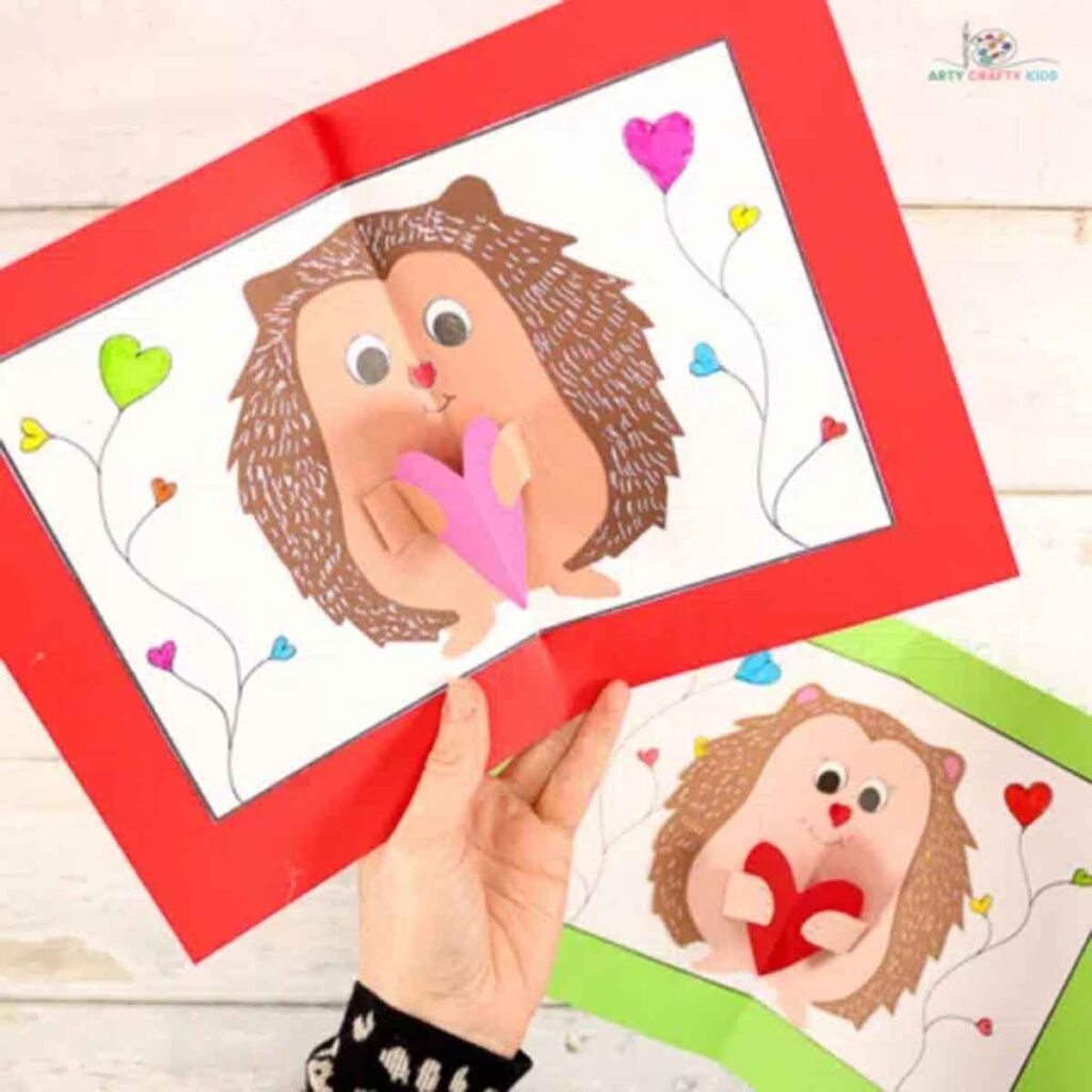 A hand presents two handmade greeting cards, perfect for Valentine paper crafts, featuring pop-up hedgehogs with colorful hearts. The cards boast red and green borders and heart-shaped balloons in vibrant shades against a rustic wooden backdrop.