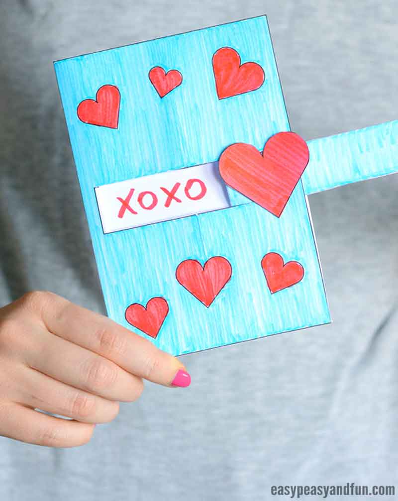 A person holds a handmade blue card adorned with red hearts, showcasing the word "XOXO" at its center—a true testament to valentine paper crafts—with a light blue sleeve peeking through. The background fades into a blurry gray, enhancing the heartfelt creation.