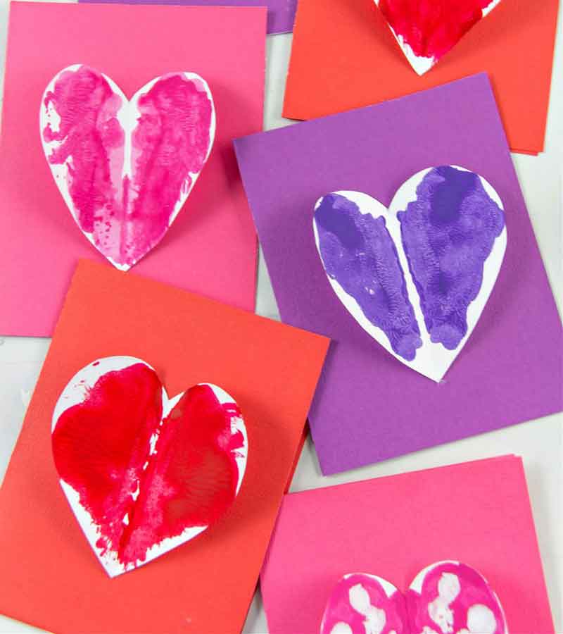 Colorful cards with heart shapes are displayed. Each heart is painted with vibrant colors including red, pink, and purple, contrasting with the solid-colored backgrounds of red and purple cards. The hearts appear to be made with folded paint techniques.