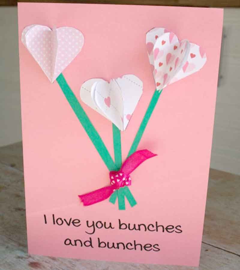 A pink handmade card with three heart-shaped paper flowers and green stems. Each flower features heart patterns. A pink ribbon is tied at the base. The text reads, "I love you bunches and bunches.