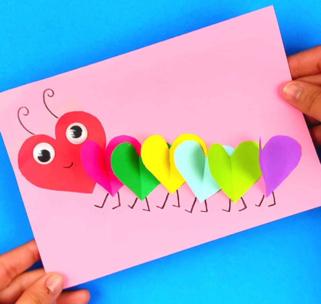 A colorful caterpillar craft made with heart-shaped paper pieces on a pink card. Each segment is a different color, with a red heart as the head. Googly eyes and cute antennae add character. Hands hold the card over a blue surface.