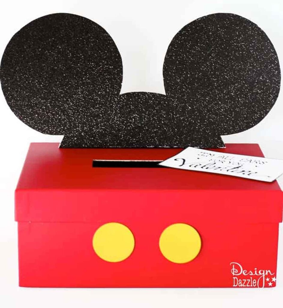 A creative valentine box idea for school, this charming red box resembles Mickey Mouse's pants with two yellow buttons on the front. Topped with large black glittery Mickey Mouse ears and a card slot, it's decorated with a small card titled "Valentine.