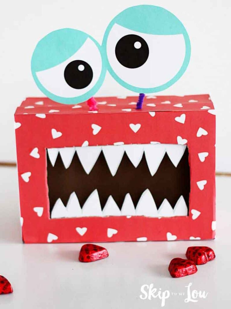 A red box decorated with white hearts embodies a charming monster theme, perfect for valentine box ideas for school. It features a large open mouth with sharp teeth and two googly eyes on top. Small heart-shaped objects are scattered around, adding a playful touch.