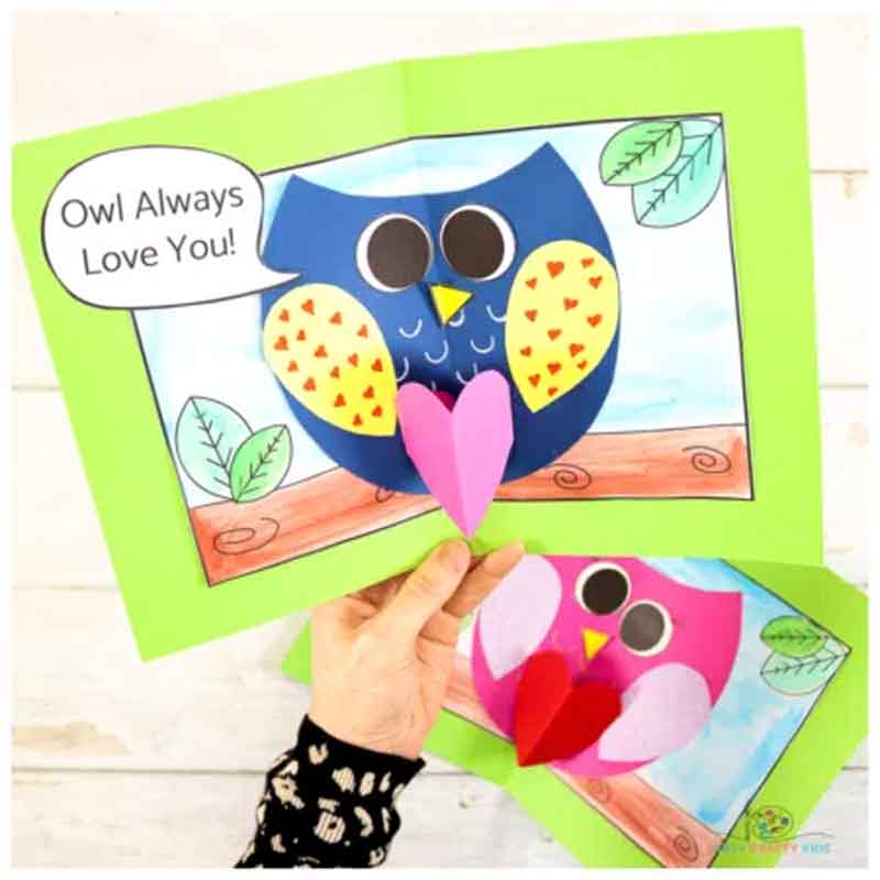 A handmade card featuring two colorful owls with heart shapes on their chests. The owls are blue and pink, set against a nature-themed background inside a green card. A speech bubble says, "Owl Always Love You!" A person's hand holds the card.