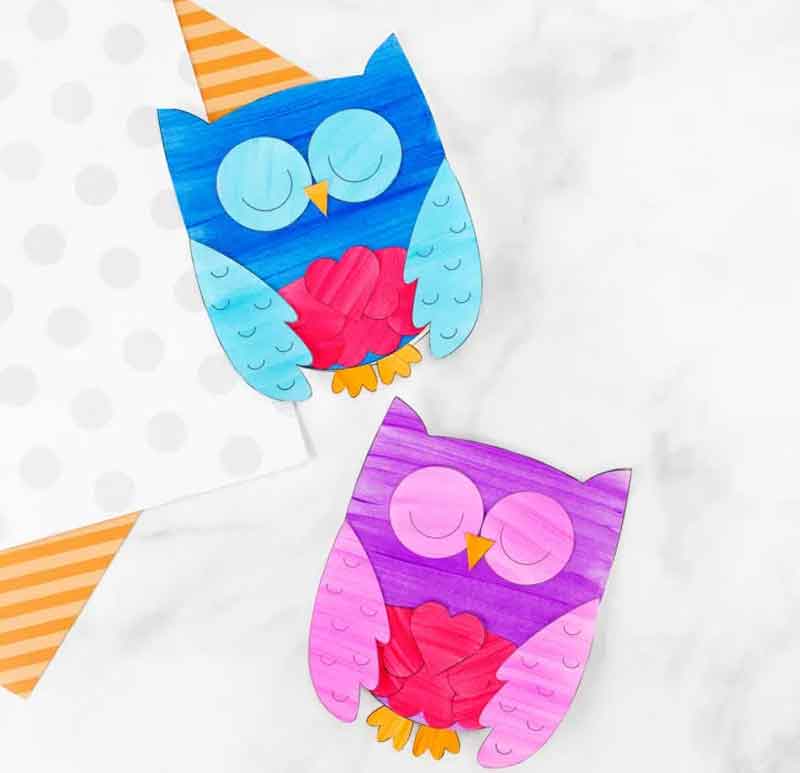 Two colorful paper owl crafts on a light background. The top owl is blue and turquoise with a heart and wears a party hat. The bottom owl is purple and pink with a heart. Both have closed eyes. Polka dot paper is partially visible.