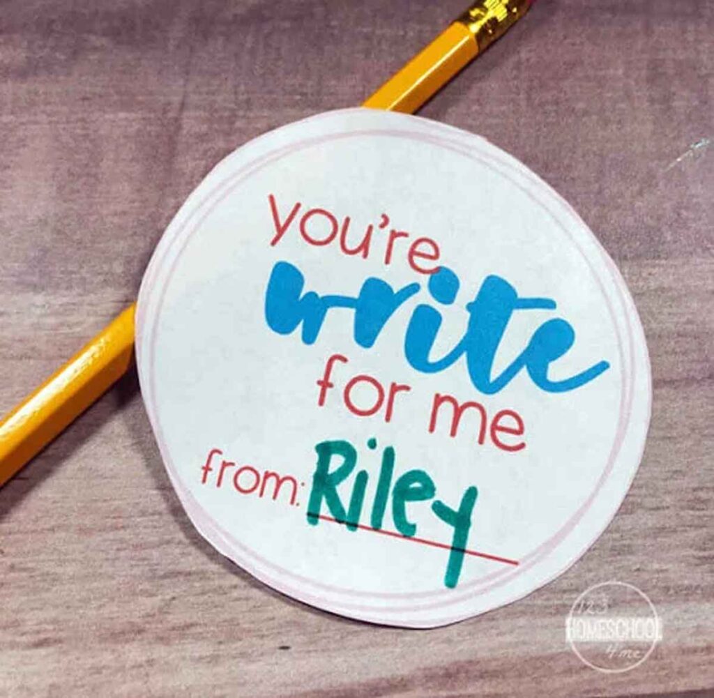 A round sticker with the text "You're write for me" in blue and red resembles a clever pencil valentine. Below, it reads "from: Riley" in green. A yellow pencil is charmingly placed next to the sticker on a wooden surface.