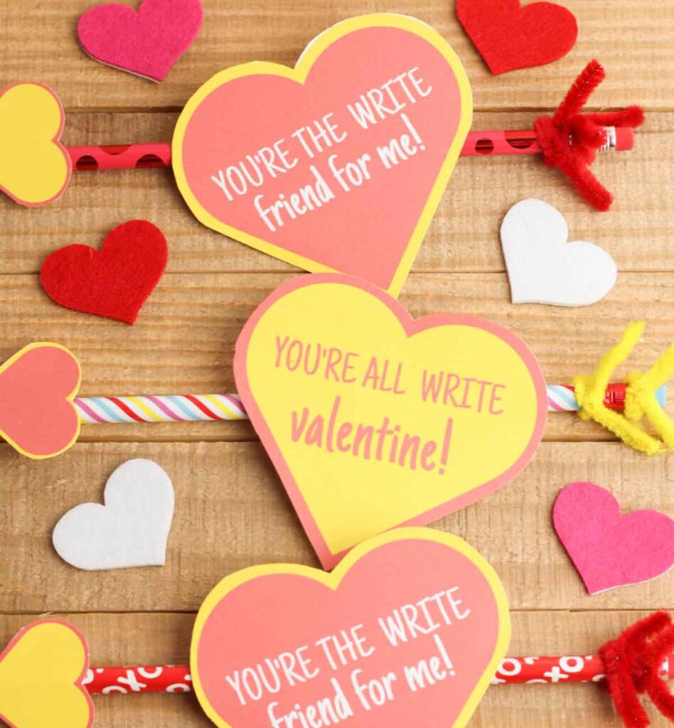 Three heart-shaped cards with puns like "You're the write friend for me!" form delightful pencil valentines. The hearts are attached to pencils and surrounded by smaller red, white, and pink paper hearts on a wooden background.