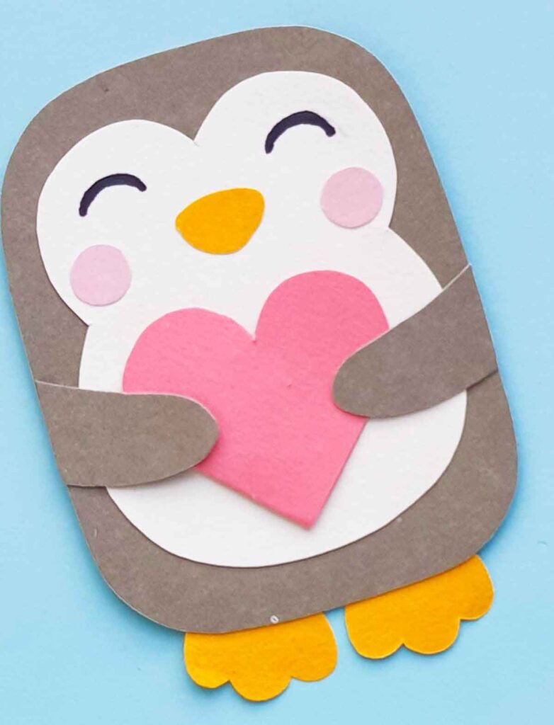 A cute valentine paper penguin craft with a gray body, white face, and orange beak and feet, holding a pink heart on a light blue background. The penguin has rosy cheeks and an adorable smile, perfect for spreading love through charming paper crafts.