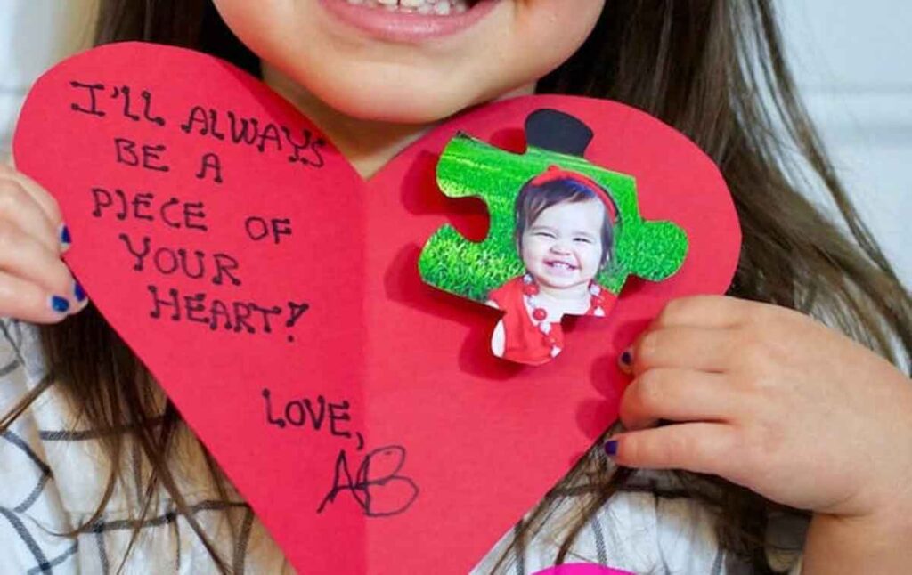 A child holds a red paper heart with handwriting that reads, "I'll always be a piece of your heart! Love, A." Attached is a green glitter puzzle piece with a photo of a smiling child. The child has painted nails and wears a striped shirt.