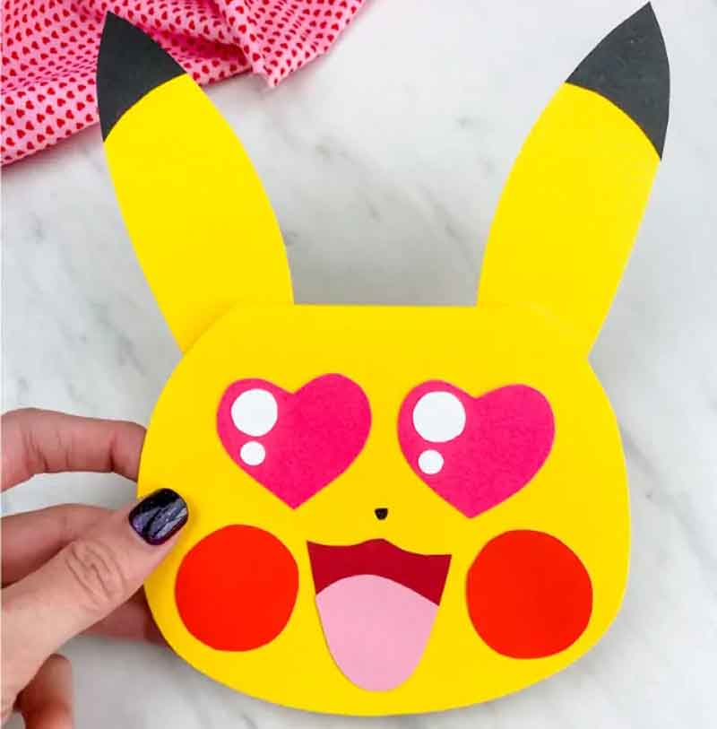 A handmade Pikachu card with heart-shaped eyes, red cheeks, and an open mouth. It is held by a person with dark nail polish. A red polka dot fabric is partially visible in the background.