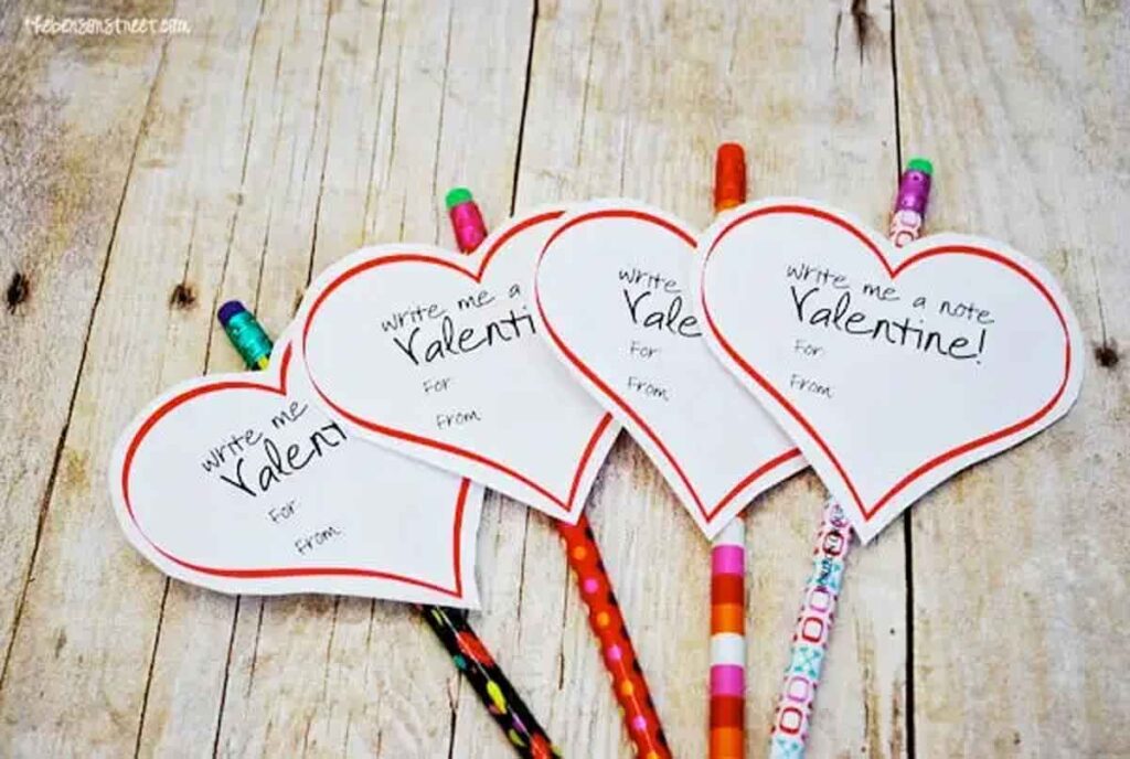 Four pencil valentines with colorful patterns are attached to heart-shaped paper reading "Write me a note Valentine!" Each heart provides spaces for "To" and "From," all charmingly arranged on a rustic wooden surface.