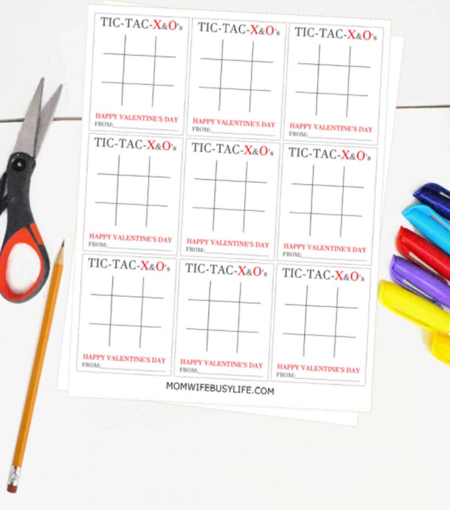 A sheet of printable tic-tac-toe Valentine's cards with red and black text, surrounded by a pair of scissors, pencil valentines, and colorful markers. The cards say "Tic-Tac-X & O's" and "Happy Valentine's Day From.