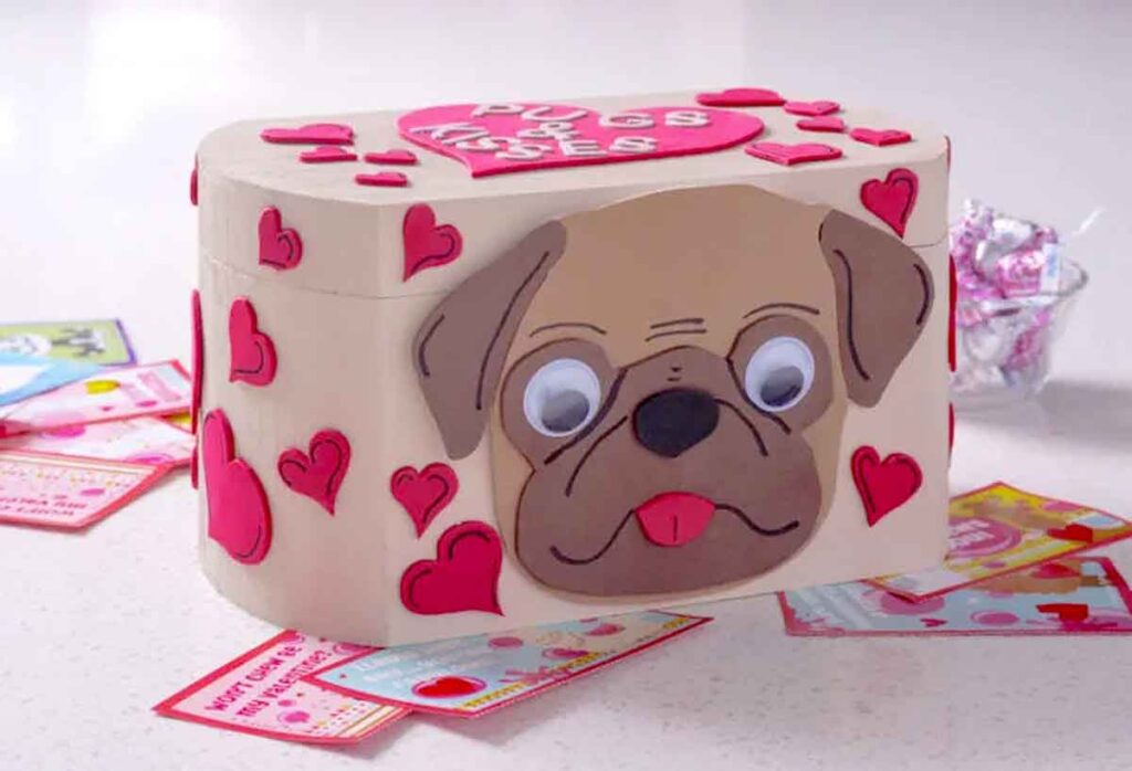 A beige and pink Valentine box with a cute dog face, googly eyes, and a small tongue. The box is adorned with red and pink hearts and stickers, displaying "pugs & kisses" on top. Perfect for Valentine box ideas for school, surrounded by Valentine's cards and candy.