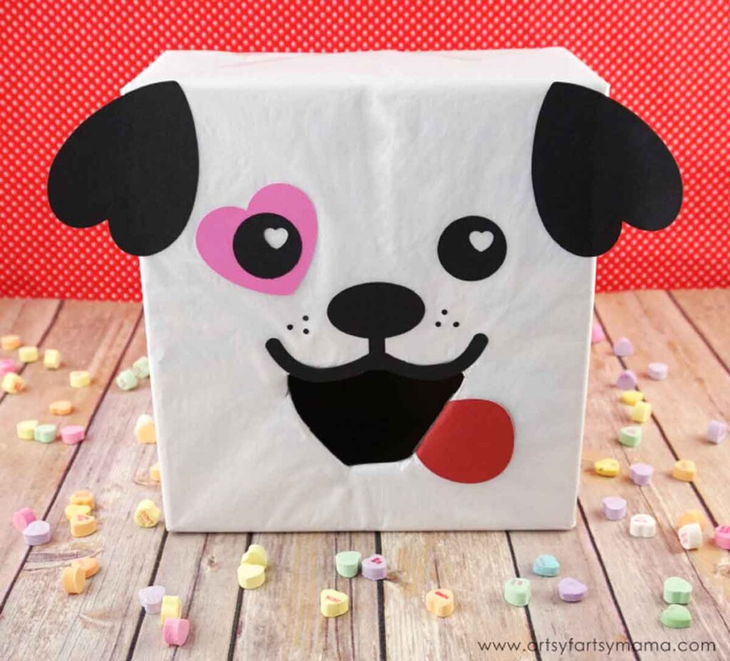 A decorated Valentine's Day box shaped like a dog, featuring black ears, a heart-shaped pink patch around one eye, and a red tongue sticking out. Perfect for school projects, it sits on a wooden surface scattered with colorful heart-shaped candies. A delightful Valentine box idea for school!.