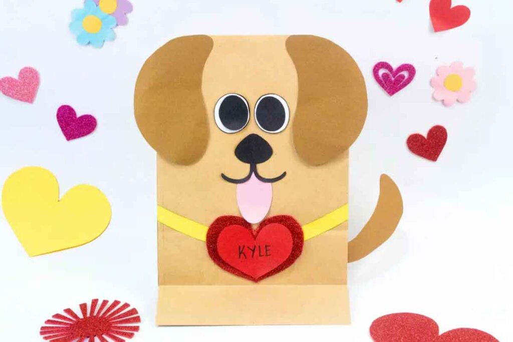 A cute Valentine paper bag puppet resembling a dog with googly eyes, a pink tongue, and heart-shaped decorations. The name "Kyle" is written on one of the hearts. Colorful hearts and flowers surround this charming piece of Valentine paper crafts.