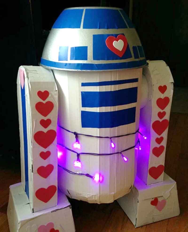 This robot costume, perfect for Valentine box ideas for school, resembles a popular sci-fi character. It's decorated with red hearts and wrapped in purple string lights. The predominantly blue and white body features silver accents, all showcased indoors on a wooden floor.