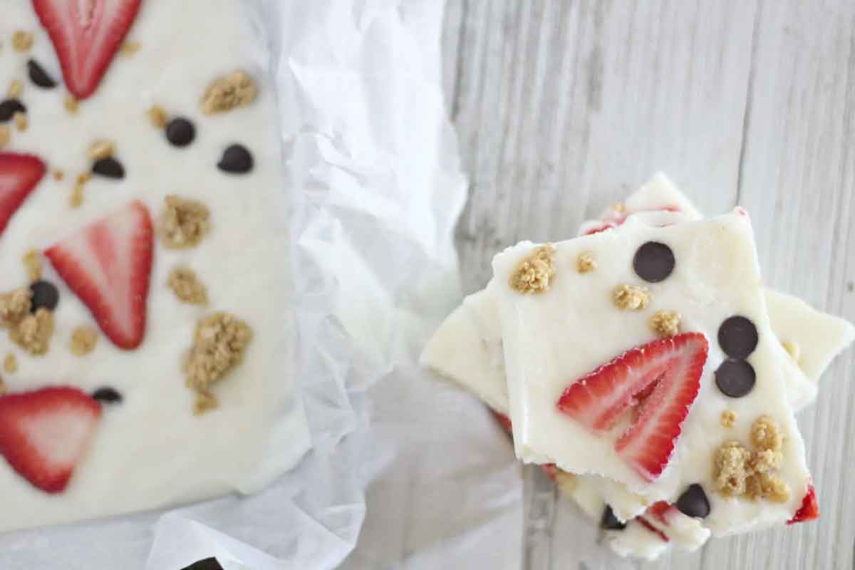 A tray of frozen yogurt bark with strawberries, chocolate chips, and granola sits on a wooden surface. Perfect as healthy Valentine snacks for the classroom, several pieces are stacked on the side, showing a close-up of the delightful toppings.