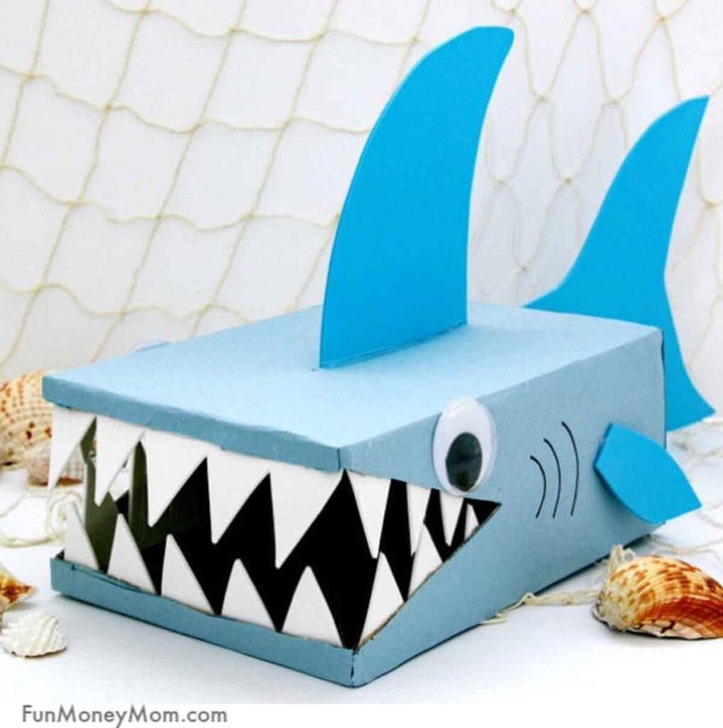 This charming valentine box idea for school is a decorative shark design featuring a blue body, large pointed teeth, and prominent fins. Complete with googly eyes, it's set against a fishnet backdrop adorned with scattered seashells.