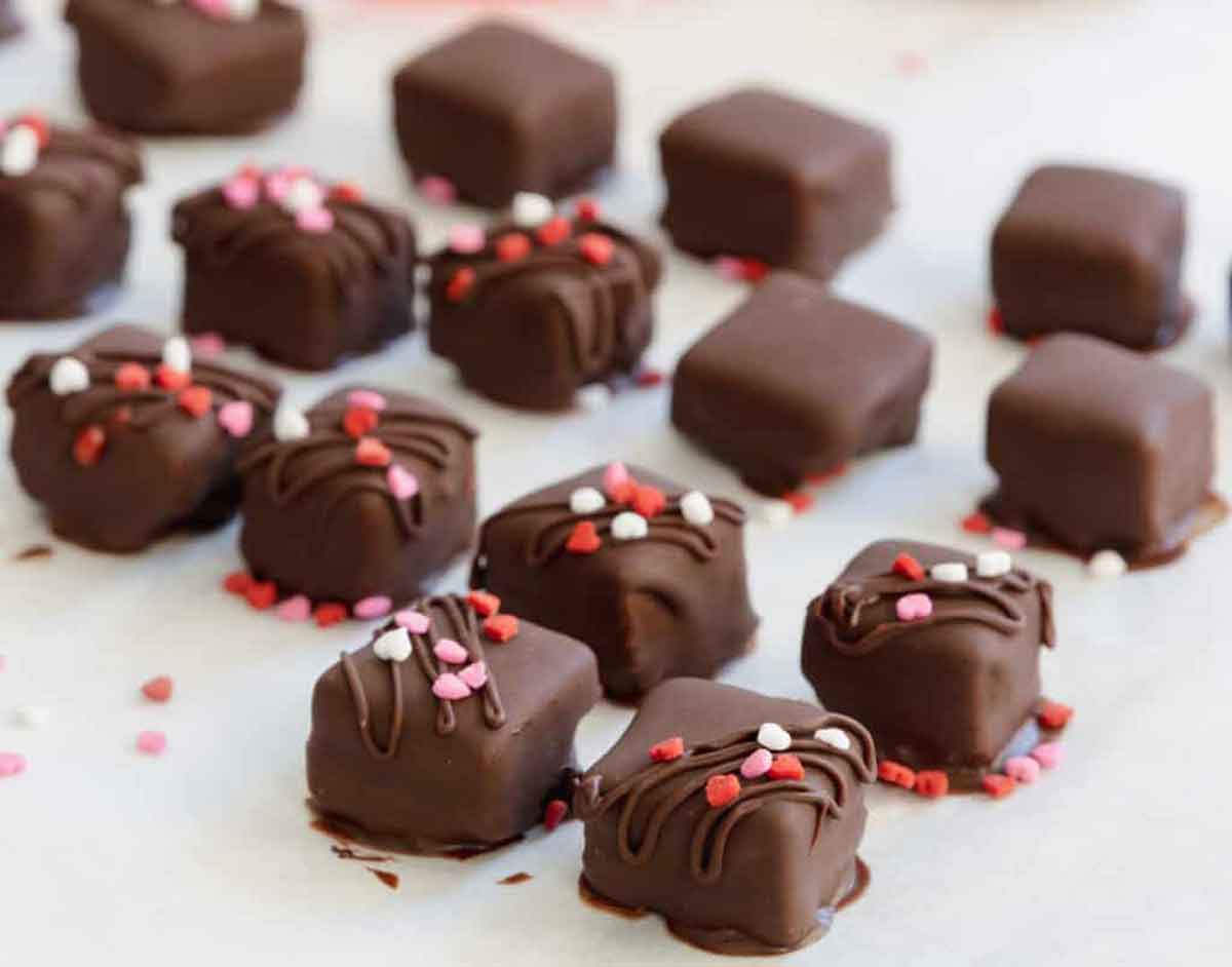 A selection of chocolate-covered square treats, perfect as healthy Valentine snacks for the classroom, are decorated with pink, red, and white sprinkles on parchment paper. Some have drizzled chocolate on top, adding a delightful textured appearance.