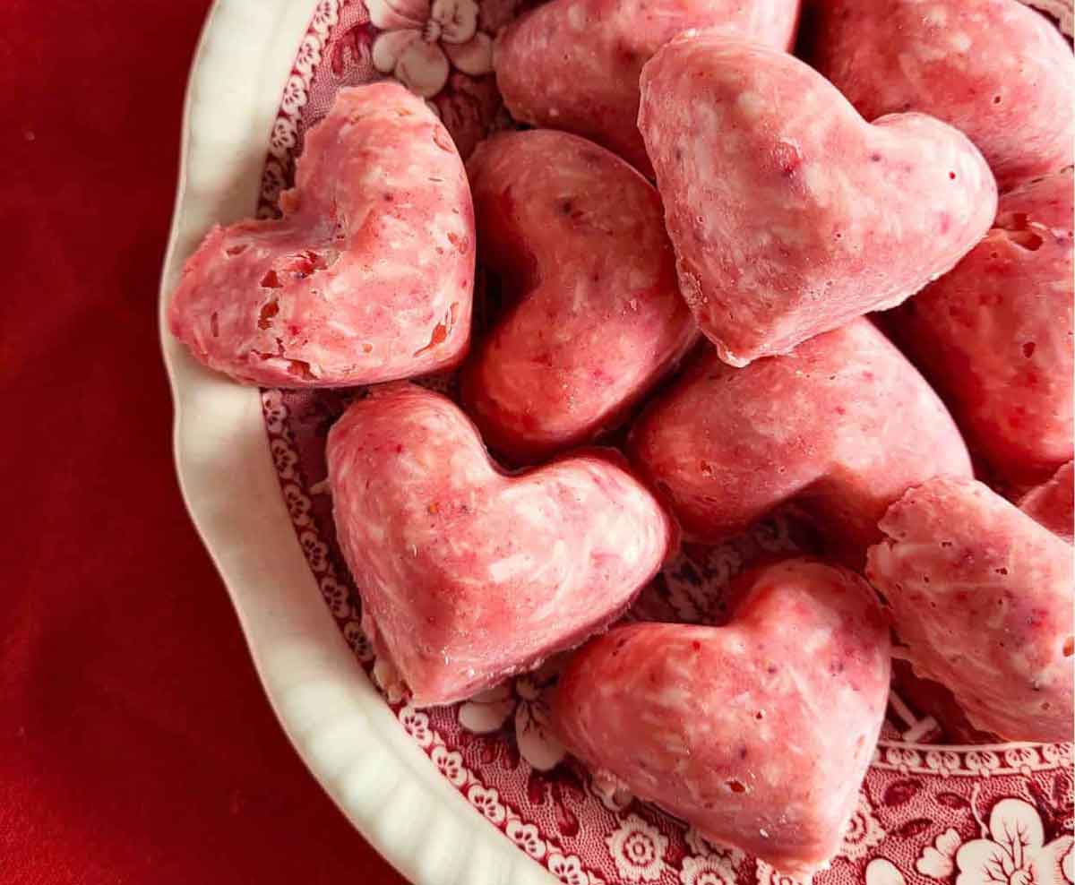Heart-shaped pink treats, perfect as healthy Valentine snacks for the classroom, are arranged in a vintage-style, floral-patterned bowl on a red surface.