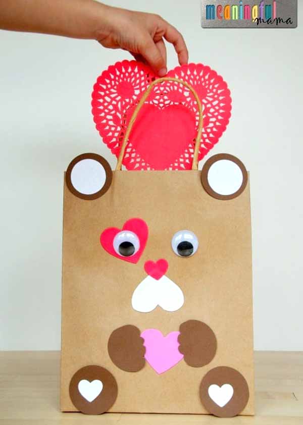 A handmade brown paper bag decorated to resemble a bear, perfect for Valentine box ideas for school. It features googly eyes and heart-shaped pink, red, and white cutouts for the ears, nose, and paws. A red heart doily is ready to be placed inside.