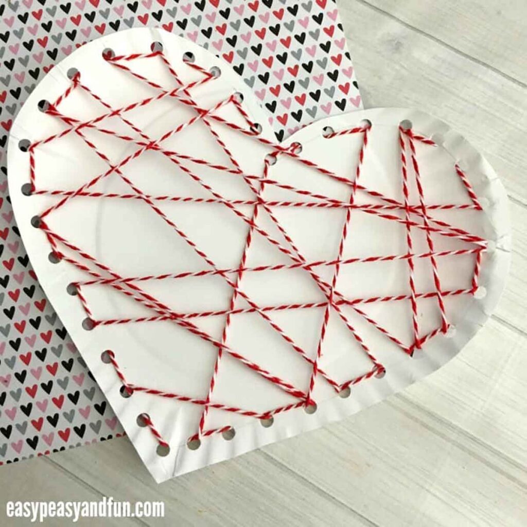 A charming paper heart, a testament to valentine paper crafts, features red and white yarn threaded through its edges in a crisscross pattern. It rests on a backdrop adorned with small, colorful hearts, capturing the spirit of love and creativity.