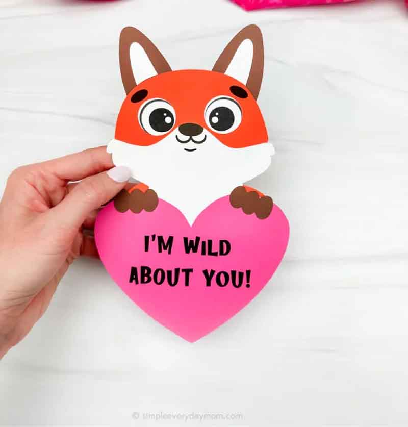 A hand holds a cute fox-shaped Valentine card, perfect for those who love valentine paper crafts. With a red face and white chest, this charming fox clutches a pink heart that reads "I'M WILD ABOUT YOU!" against a crisp white background.