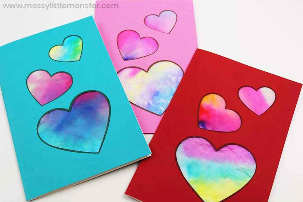 Three colorful handmade cards with heart cutouts. The hearts reveal a vibrant, watercolor-like background blending pink, blue, and yellow hues. The cards are in blue, pink, and red.