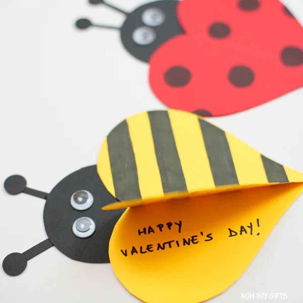 A Valentine's Day craft with a ladybug and a bee. The bee, with yellow and black stripes, has heart-shaped wings. The open wing displays the message "HAPPY VALENTINE'S DAY!" The ladybug has red and black heart-shaped wings.
