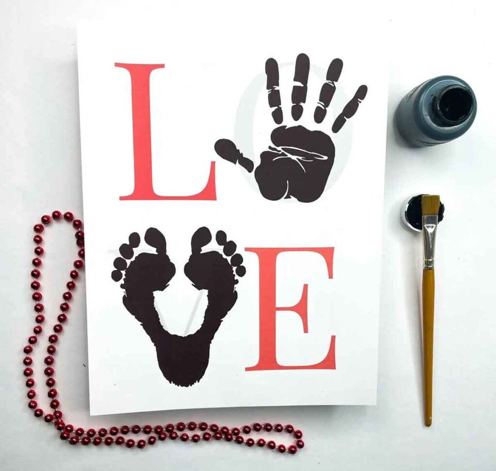 A creative Valentine paper craft artwork spells "LOVE" with a red "L" and "E", while black hand and footprints form the "O" and "V". Nearby, a red beaded necklace, paintbrush, and paint jar rest on a white background.
