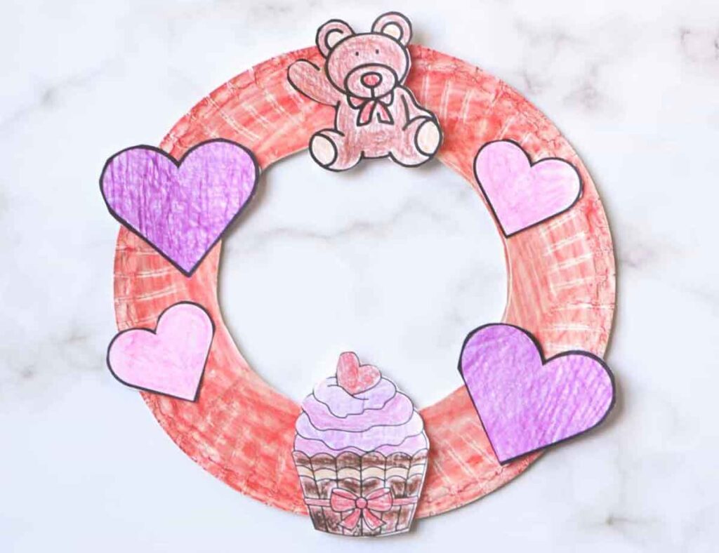 A delightful Valentine's paper craft, this wreath is crafted from a red paper plate and features a charming teddy bear, three purple hearts, and a cupcake with pink frosting and a cherry on top, all elegantly displayed on a marble background.