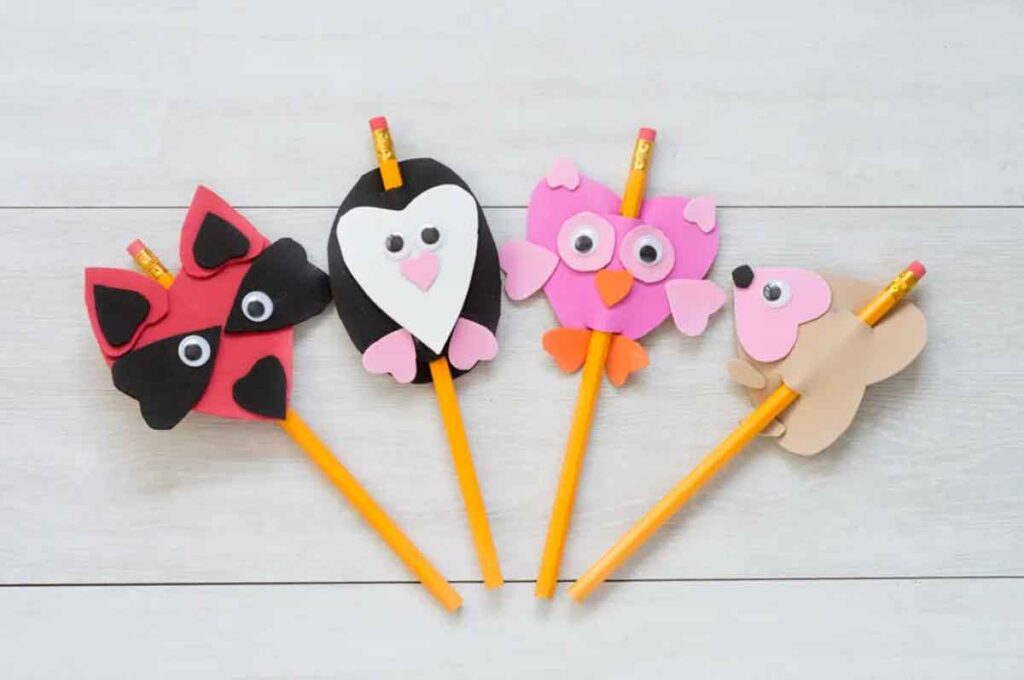 Four pencils on a white wooden surface, each topped with a colorful animal-shaped felt decoration: a fox, a penguin, an owl, and a bear. These charming pencil valentines wrap their soft bodies around the center of the pencil.