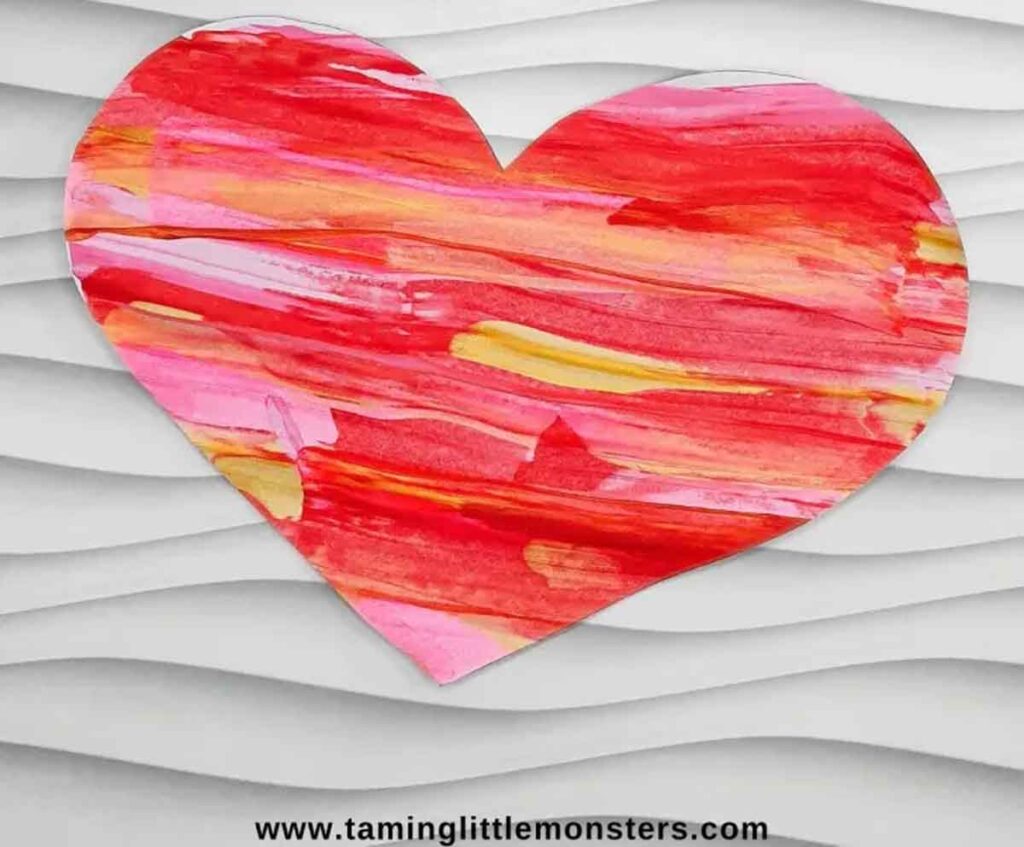 A vibrant heart-shaped paper featuring red, pink, orange, and yellow brushstrokes on a textured white background, perfect for Valentine paper crafts. Website URL "www.taminglittlemonsters.com" is visible at the bottom.