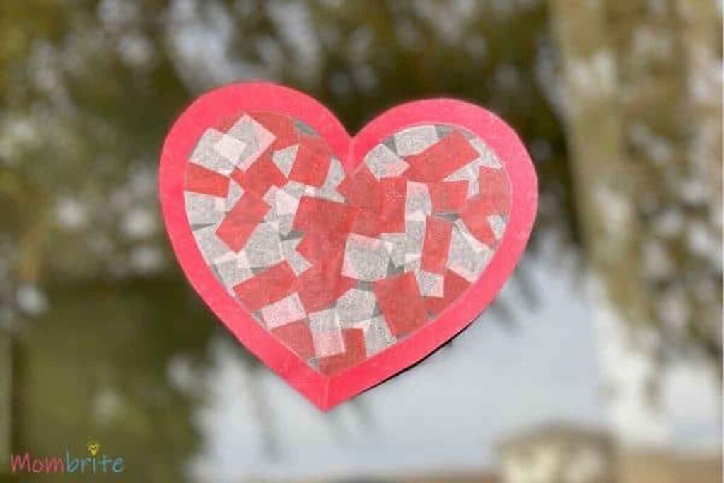 A handcrafted heart with a red outline and a mosaic of red and white tissue paper pieces is displayed on a glass window, embodying the charm of Valentine paper crafts, with blurred greenery visible in the background.