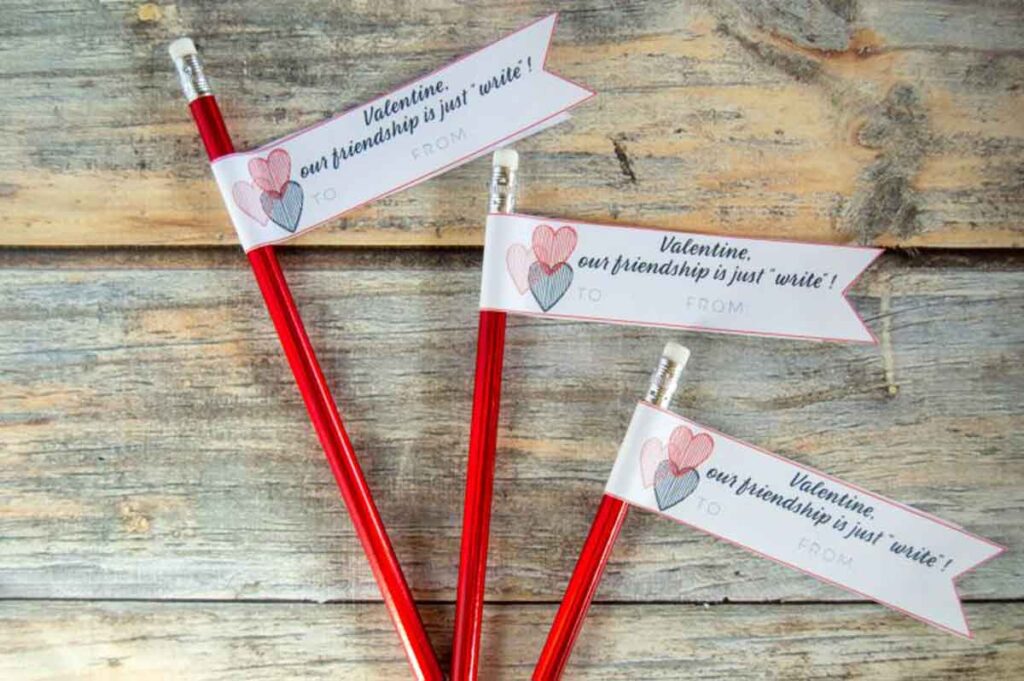 Three red pencil valentines with erasers are attached to paper banners that read, "Valentine, our friendship is just 'write'," adorned with heart designs. The banners have spaces labeled "TO" and "FROM," and the pencils rest on a rustic wooden surface.