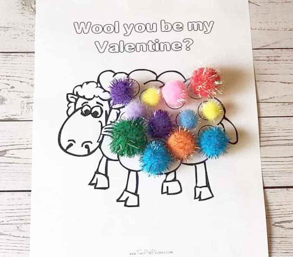 A coloring sheet with a cartoon sheep, with colorful pom-poms glued onto its body, resembling wool. The text above the sheep reads, "Wool you be my Valentine?" The background is a wooden surface.