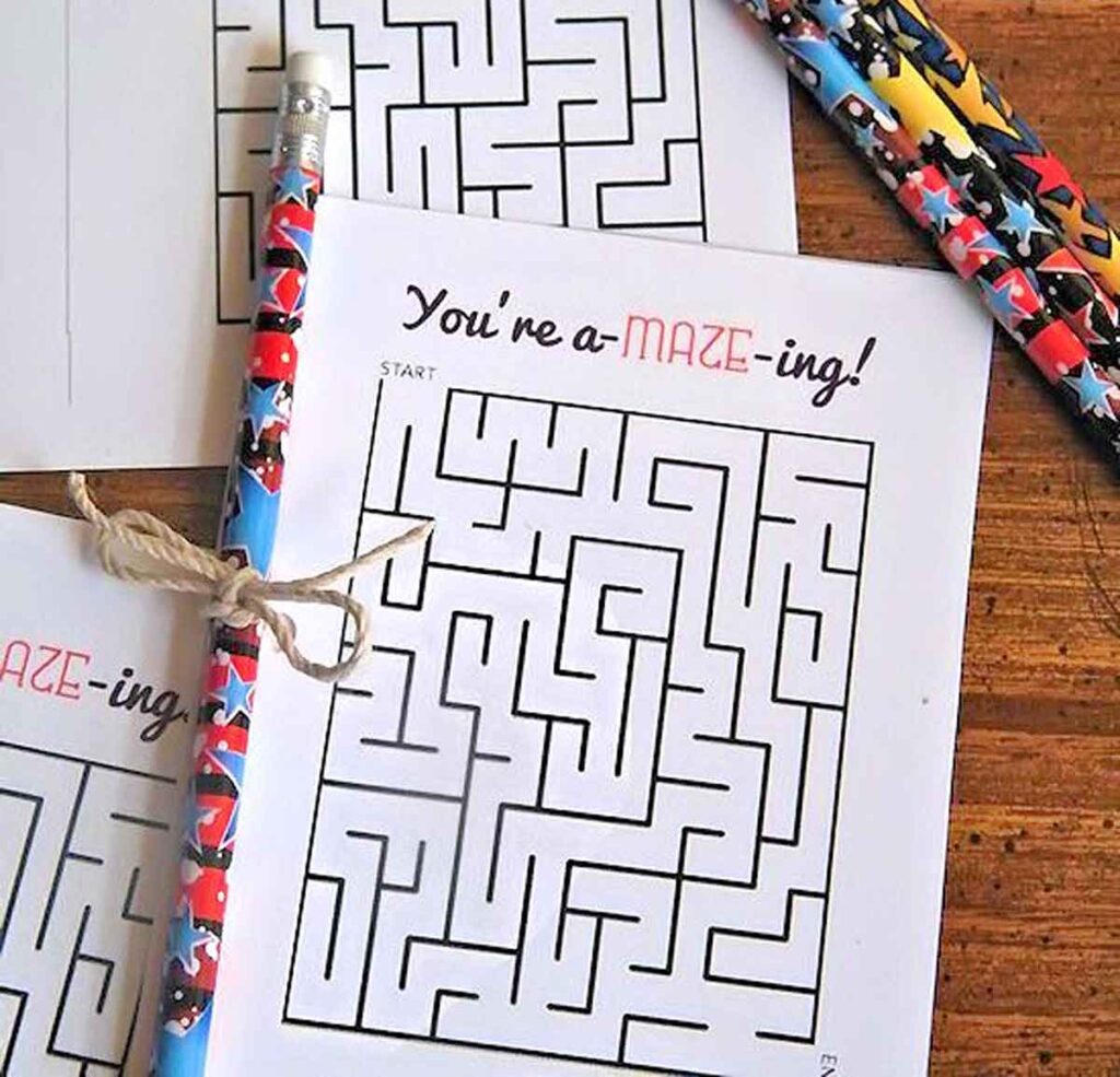 A printed maze puzzle with the words "You're a-MAZE-ing!" at the top and a decorated pencil valentines resting on it. Another pencil and more maze sheets are nearby on a wooden surface, creating an ambiance of playful creativity.