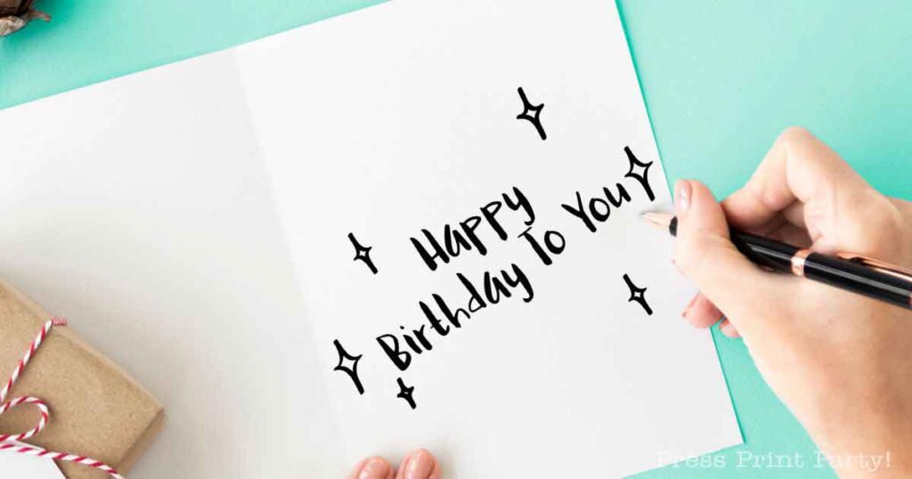 A person inscribes "Happy Birthday To You" with black ink inside an open card on a light turquoise surface. Next to it, a small wrapped gift peeks in, while sparkle doodles dance around the text, conveying the best birthday wishes.