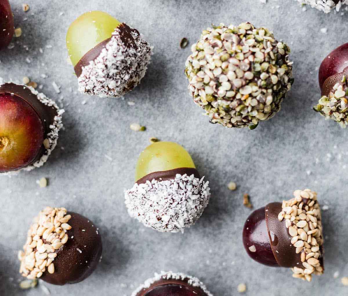 A delightful array of chocolate-covered fruits sits on parchment paper, perfect as healthy Valentine snacks for the classroom. Grapes and small red fruits are adorned with toppings like shredded coconut, seeds, and nuts, creating a treat that's both nutritious and festive.