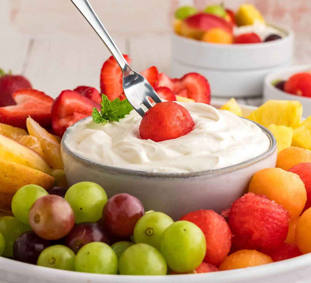 A bowl of creamy white fruit dip is the perfect healthy Valentine snack for the classroom, surrounded by fresh grapes, melon balls, strawberries, pineapple, and apple slices. A fork holds a watermelon ball above the dip for a sweet and fun touch.