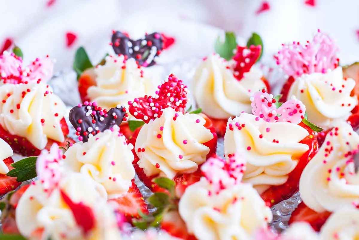Close-up of small, healthy Valentine snacks for the classroom topped with white cream, pink and red heart-shaped decorations, and sprinkles. Fresh strawberries and mint leaves are visible beneath the cream, creating a festive and colorful presentation.