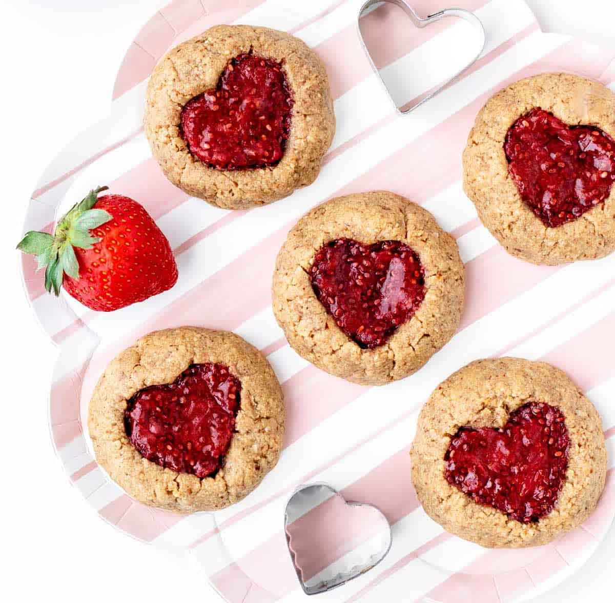 Heart-shaped cookies filled with red jam are arranged on a striped tray, perfect as healthy Valentine snacks for the classroom. A fresh strawberry and two heart-shaped cookie cutters accompany the cookies.