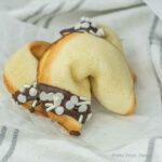 A fortune cookie dipped in chocolate and decorated with white sprinkles, placed on a piece of white parchment paper with a striped cloth underneath.