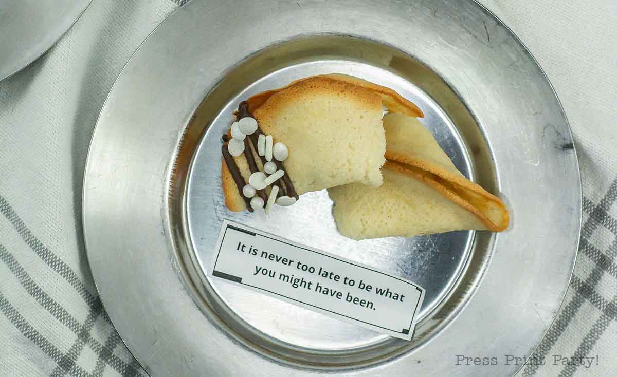 A fortune cookie with chocolate drizzle and sprinkles open on a silver plate. The fortune slip reads, "It is never too late to be what you might have been." A white and gray striped cloth is partially visible underneath.