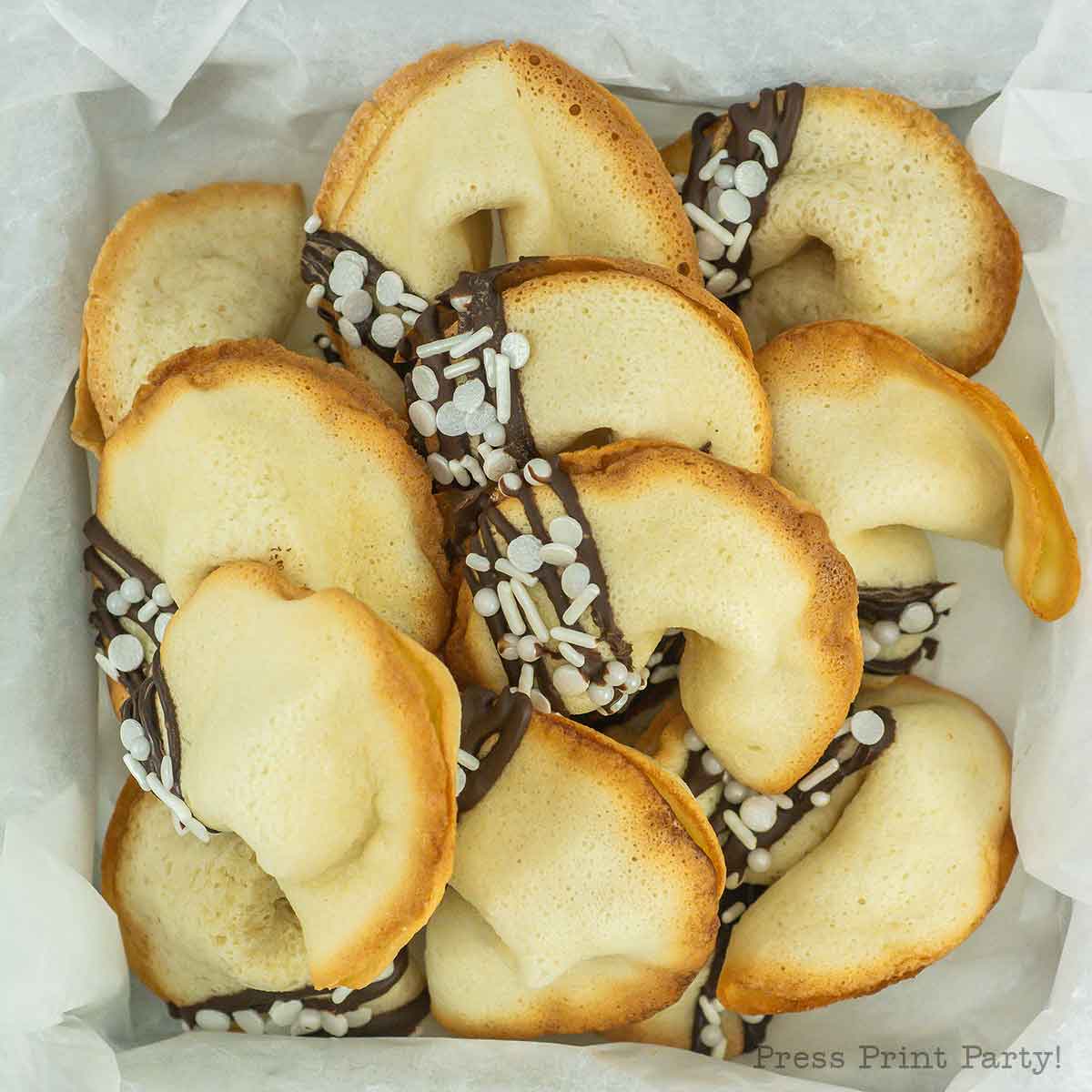 A box filled with fortune cookies partially dipped in chocolate and sprinkled with white decorations. They are arranged on white parchment paper.