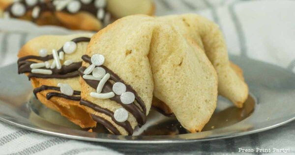A fortune cookie decorated with chocolate drizzle and silver sprinkles rests on a metal plate. The background features a striped cloth.