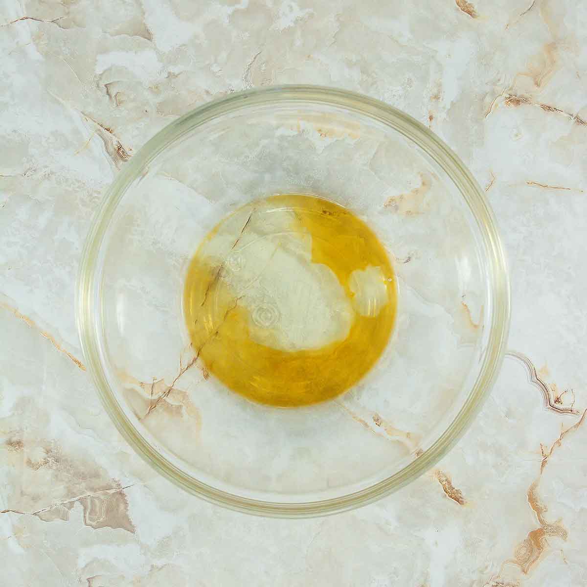 A clear glass bowl containing a mixture of wet ingredients, possibly oil and water, placed on a marble surface with light brown and beige veining.
