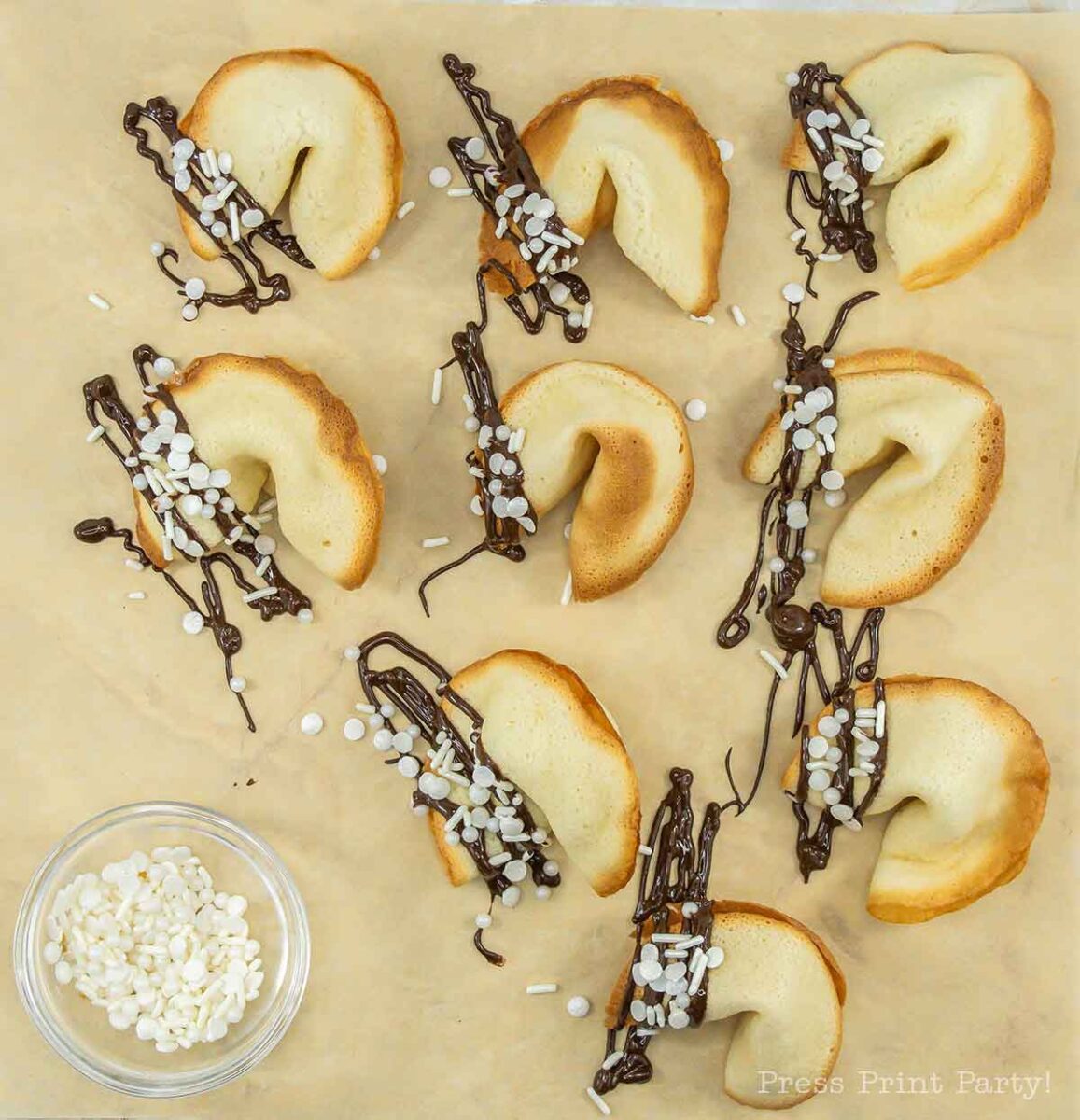 Eight fortune cookies drizzled with dark chocolate and sprinkled with white sugar pearls, arranged on parchment paper. A small bowl of sugar pearls is placed beside them.
